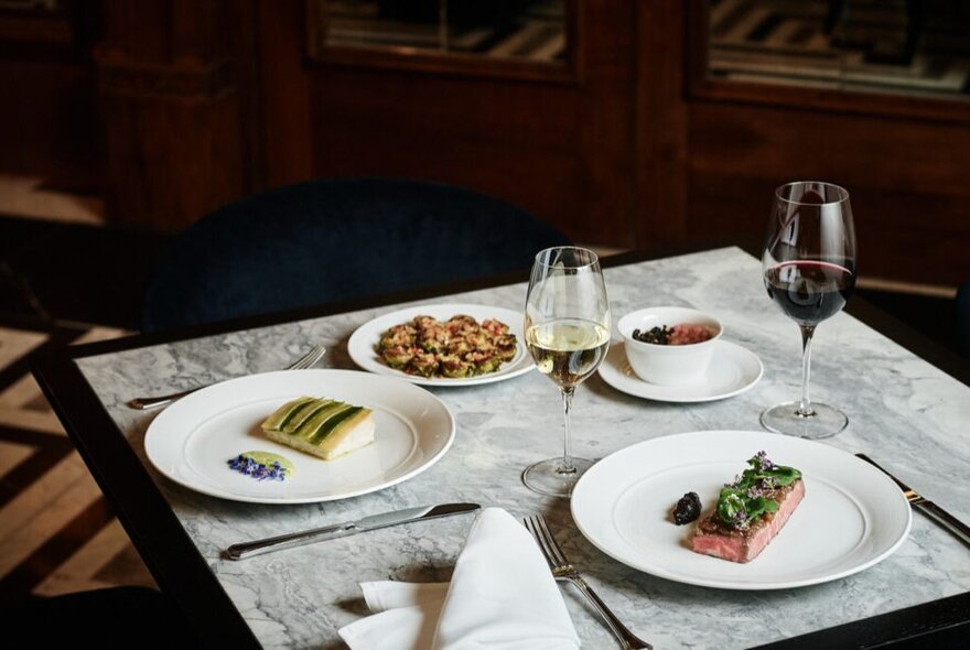 A table for two in a restaurant, with two main courses on white plates, two side dishes and two glasses of wine, napkins and cutlery.