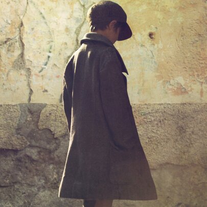 A young person wearing a cap and a long coat standing against a weathered, textured wall. 