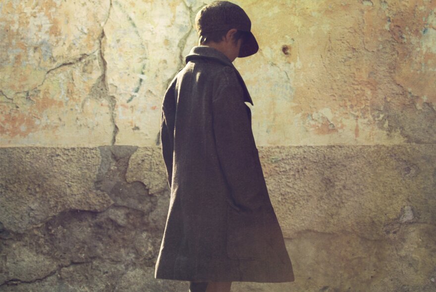 A young person wearing a cap and a long coat standing against a weathered, textured wall. 
