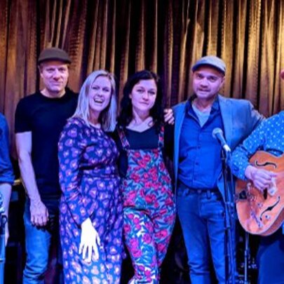 Six-piece musical group bathed in blue light, standing in front of gold curtains.