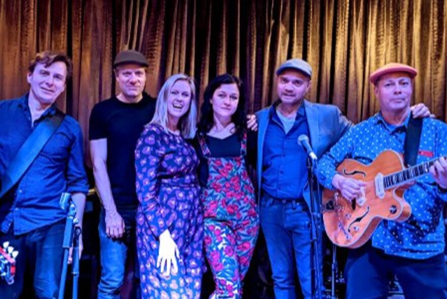 Six-piece musical group bathed in blue light, standing in front of gold curtains.