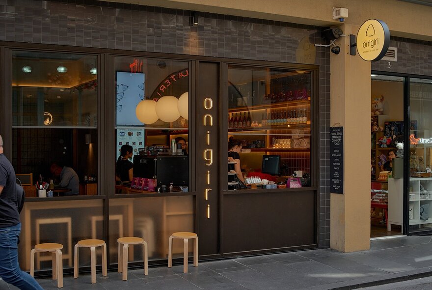 Exterior of a restaurant with small stools on the footpath facing an open window.
