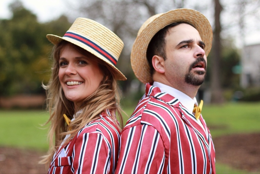 Man and woman standing back to back outside, wearing matching red, white and black striped jackets and straw boaters.