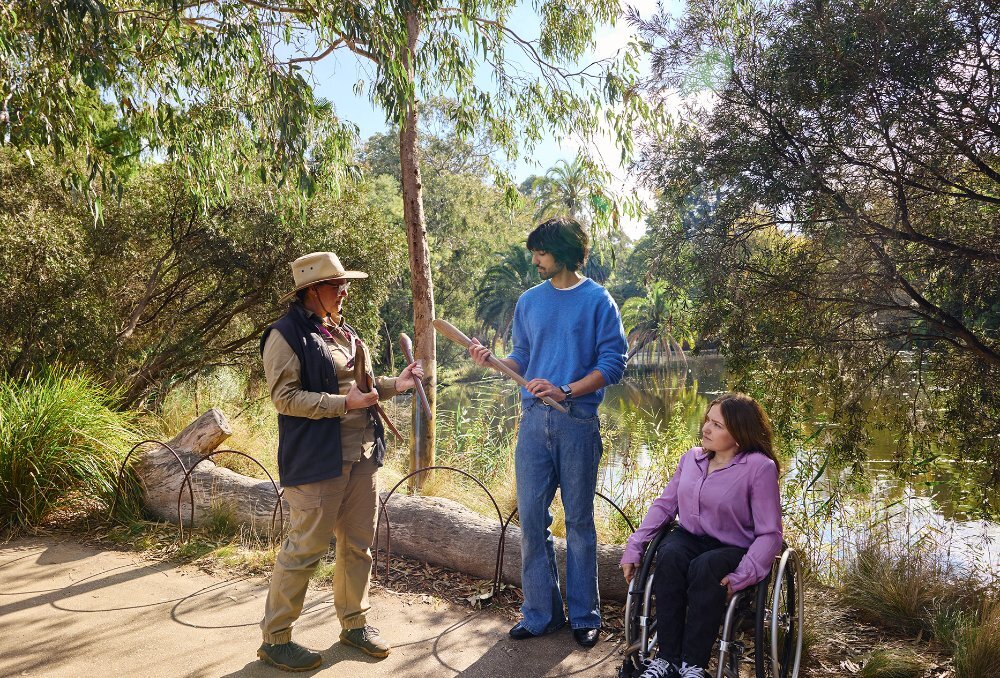 A guide in outdoor clothing chatting to two people in a garden. They are holding Aboriginal artefacts, the woman is using a wheelchair.