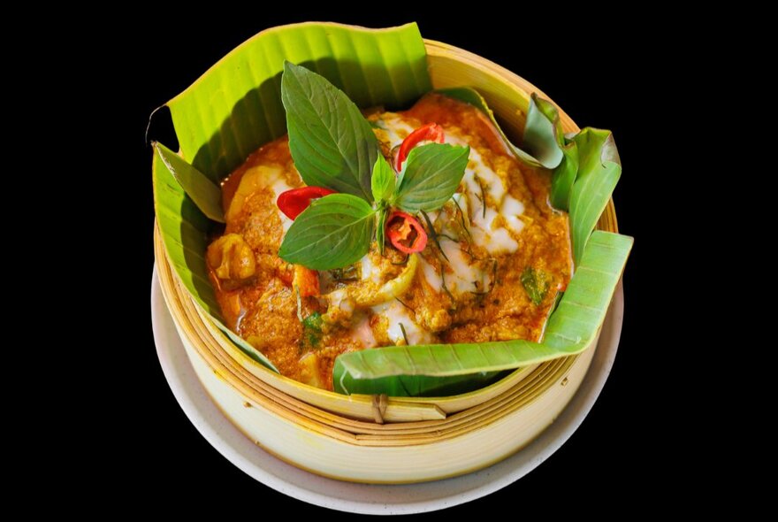 Looking down at a banana leaf lined bamboo steamer containing Cambodian food with Thai basil and chilli garnish. 