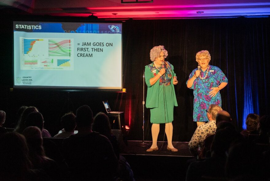 Two people performing on a small stage in front of a seated audience, a powerpoint style presentation on a large screen to the right of them.