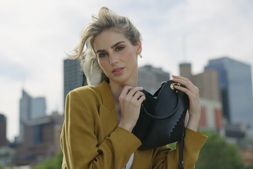 Woman, wearing mustard-coloured jacket standing against cityscape, holding black handbag up to left shoulder.