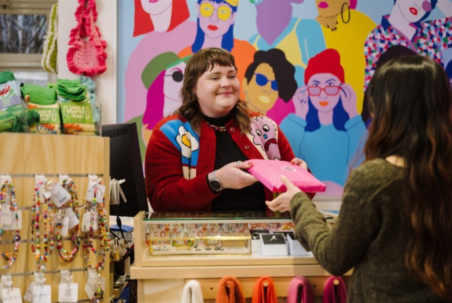 A person behind a shop counter handing a wrapped item to a shopper inside the Queen Victoria Women's Centre SHOP!