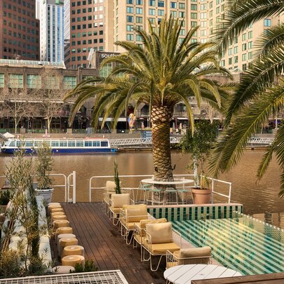 The deck of Afloat on the Yarra River, with tables and chairs, a low wading pool, palm trees and views of city buildings in the background.