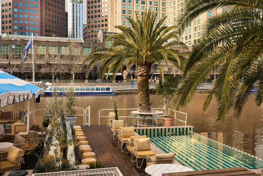 The deck of Afloat on the Yarra River, with tables and chairs, a low wading pool, palm trees and views of city buildings in the background.