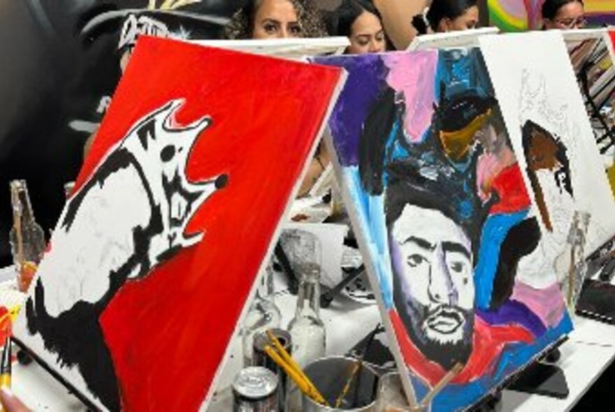 Close up of two square, half-painted canvases, other canvases and painters' faces peeking out from a row at rear.