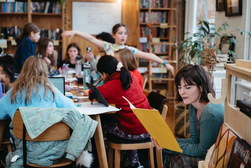 Primary school-aged children sitting, writing and interacting with each other around a table in a writing studio, an adult teacher nearby reading, bookshelves and a whiteboard on a rear wall.