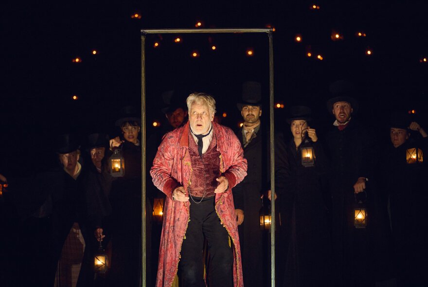 A man in a red dressing gown surrounded by characters holding torches, on stage.