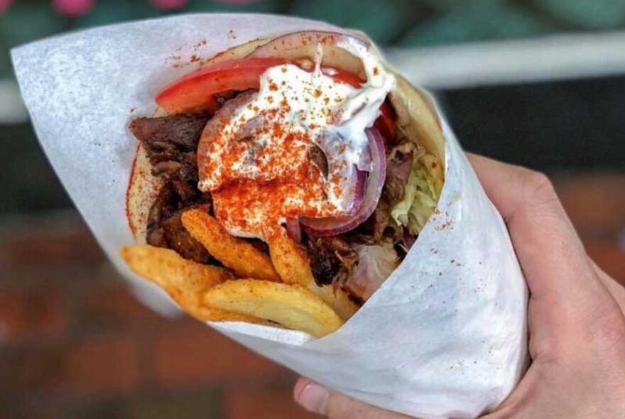 A hand holding a souvlaki with chips and tzatziki.