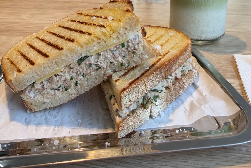 A close-up of a toasted tuna melt panini, sliced in half, resting on a white paper liner on a metallic serving tray.