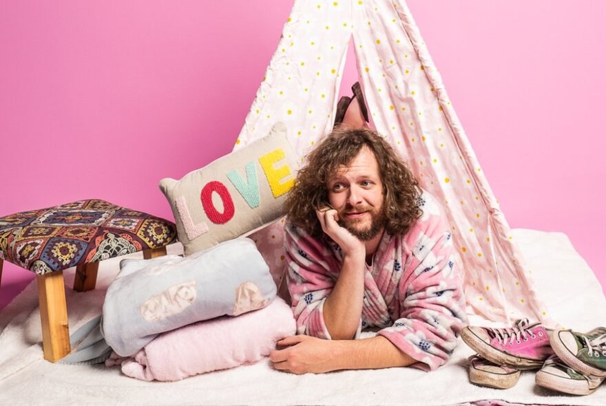 Man wearing a dressing gown, lying on the floor in a child's teepee, surrounded by cushions and a rug.