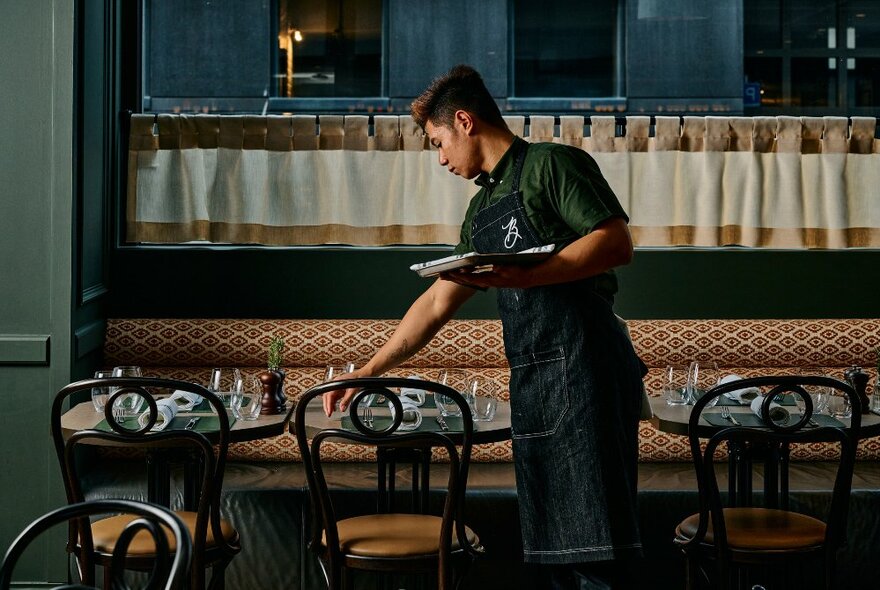 A waiter preparing a dinner service in a restaurant by placing cutlery on the tables.