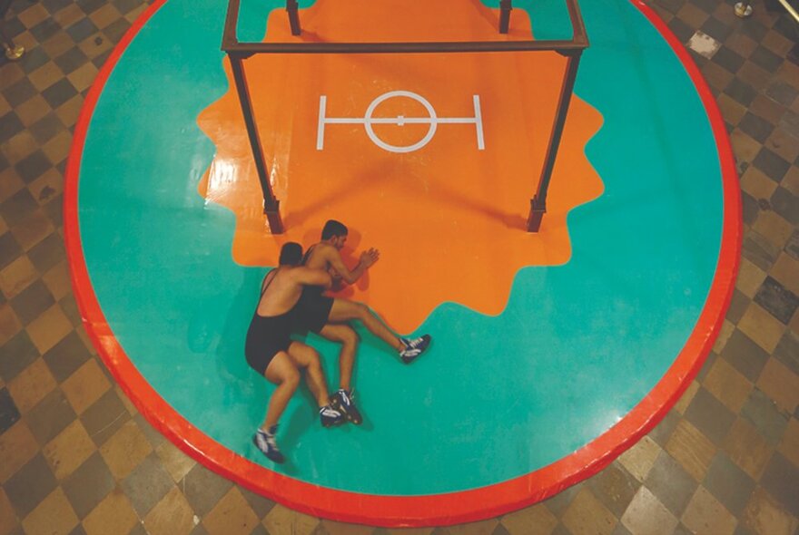 Looking down at two men wrestling on the ground inside a red rimmed circle on a jade green background.