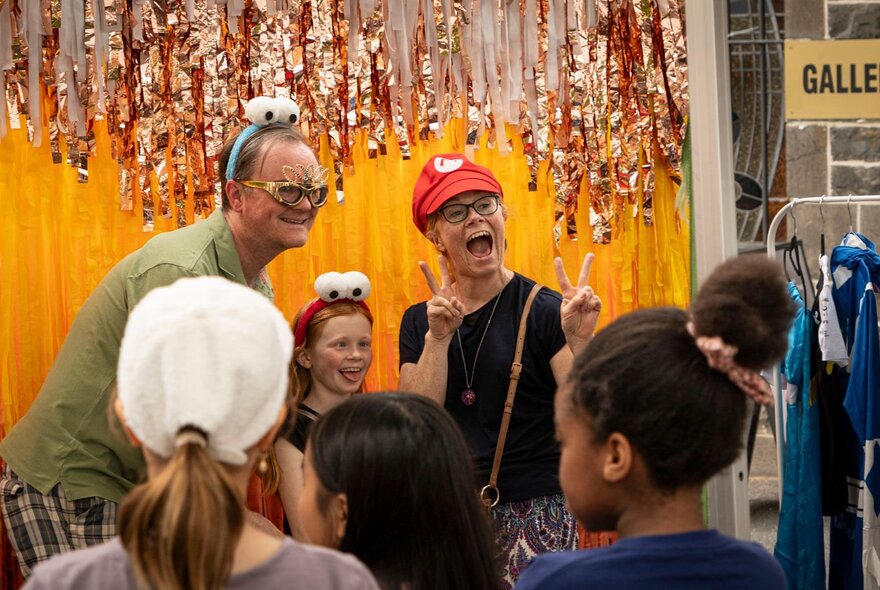 Three people having a photo taken of them with funny hats and headbands, in front of a gold and yellow streamers.