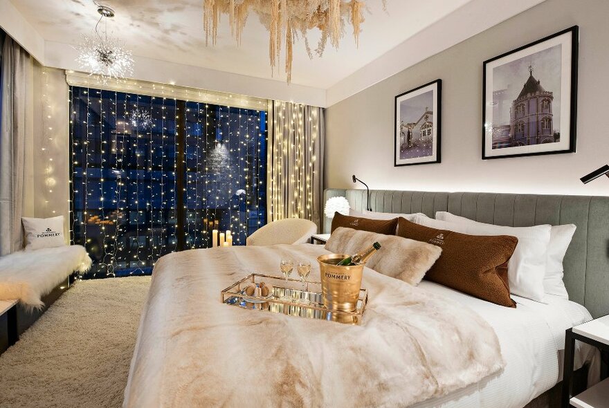 A hotel room with fairy light curtains, a furry throw and a bottle of Champagne on the bed.