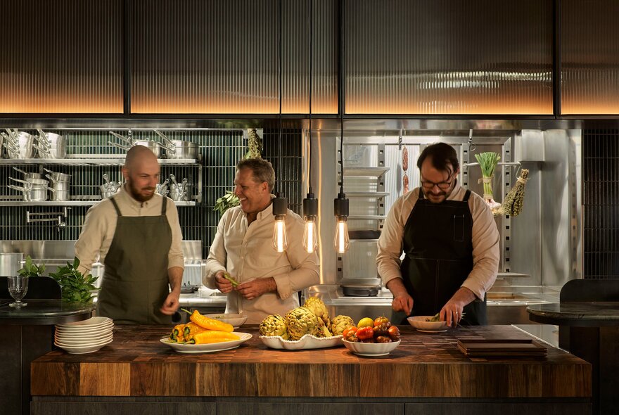 Three chefs laughing together as they prepare food in a professional kitchen.