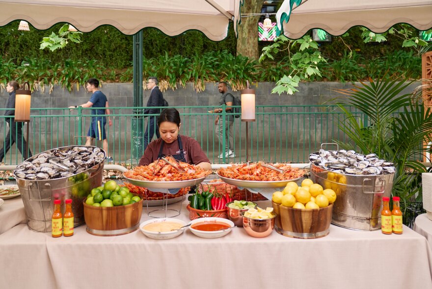 An abundant seafood buffet table at Arbory Afloat with a person preparing food behind it, city passers by in the background.