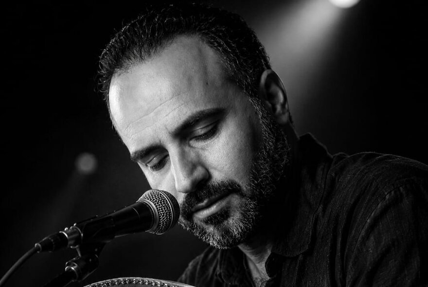 A black-and-white close-up photograph of a man with short dark hair and a beard, singing or speaking into a microphone, his face and eyes downcast.