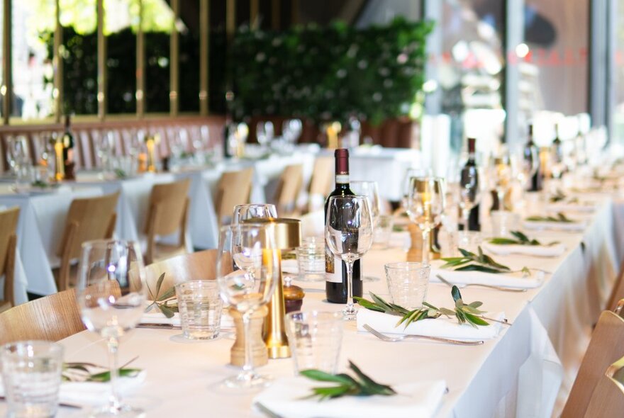 A long rectangular dining table with a white tablecloth, cloth napkins, glasses and bottle of wine, inside a restaurant.