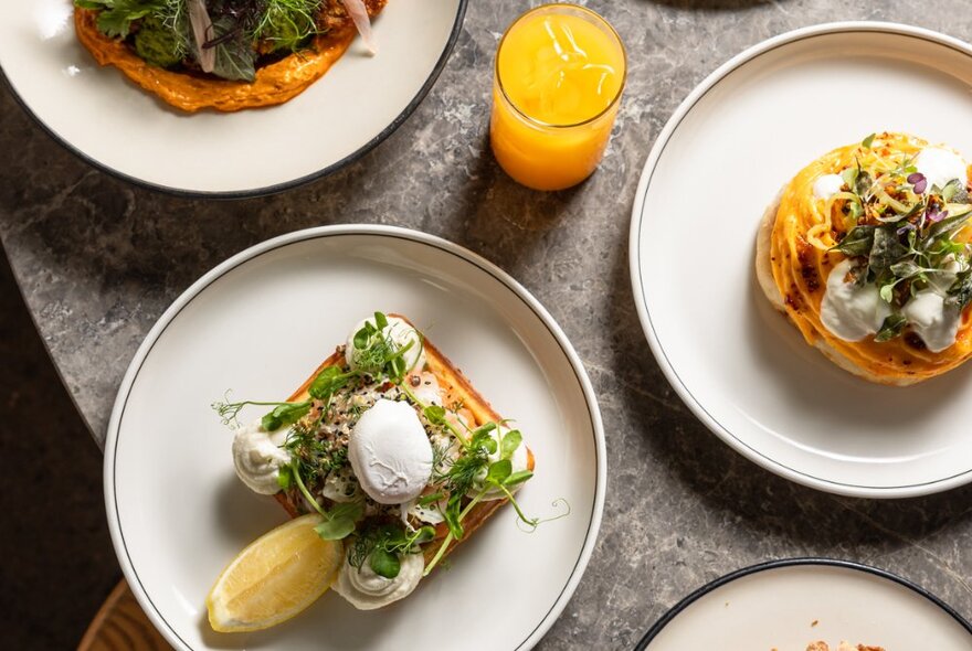 Several breakfast dishes on a concrete table, including poached eggs and orange juice.