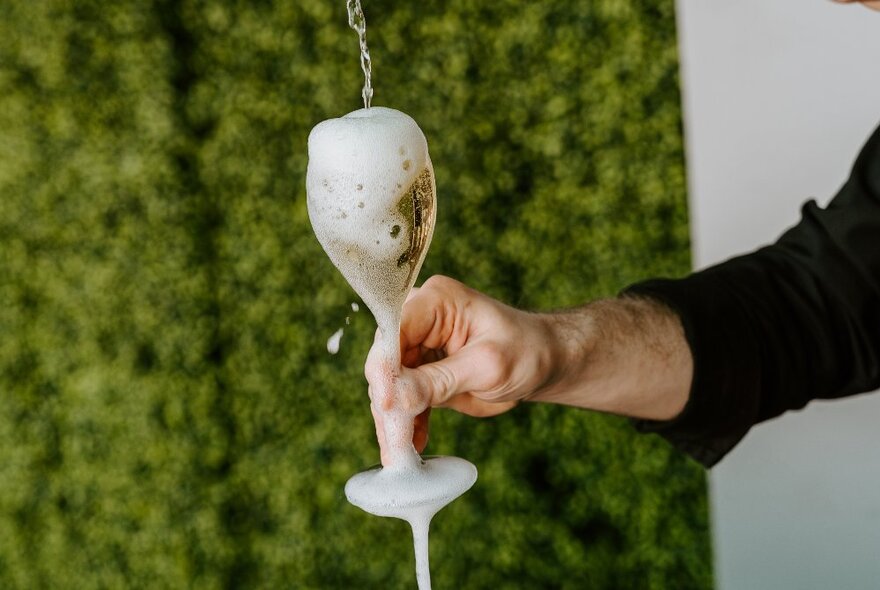 A hand holding out an overflowing flute glass of sparkling wine.