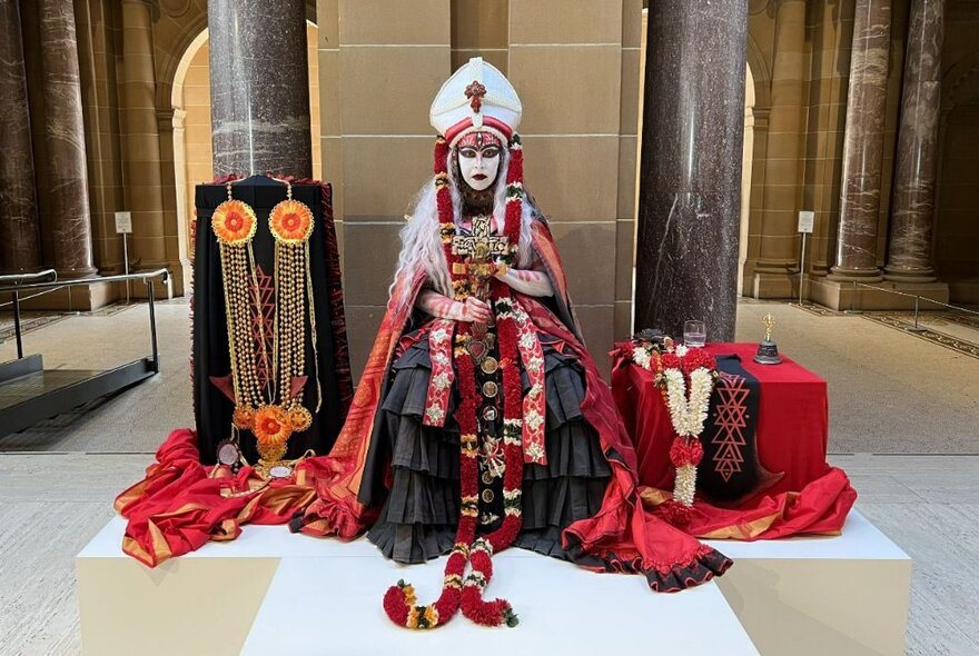 A seated performer wearing elaborate red, white and black garments, a papal headdress, garlands of flowers around her neck, in full theatrical makeup and in a spacious room featuring marble columns.