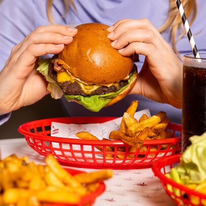 A person holding a classic cheeseburger with lettuce and a side of fries in a red basket, accompanied by a soft drink with a straw.