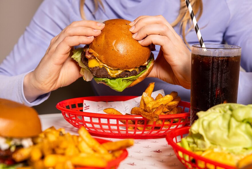 A person holding a classic cheeseburger with lettuce and a side of fries in a red basket, accompanied by a soft drink with a straw.