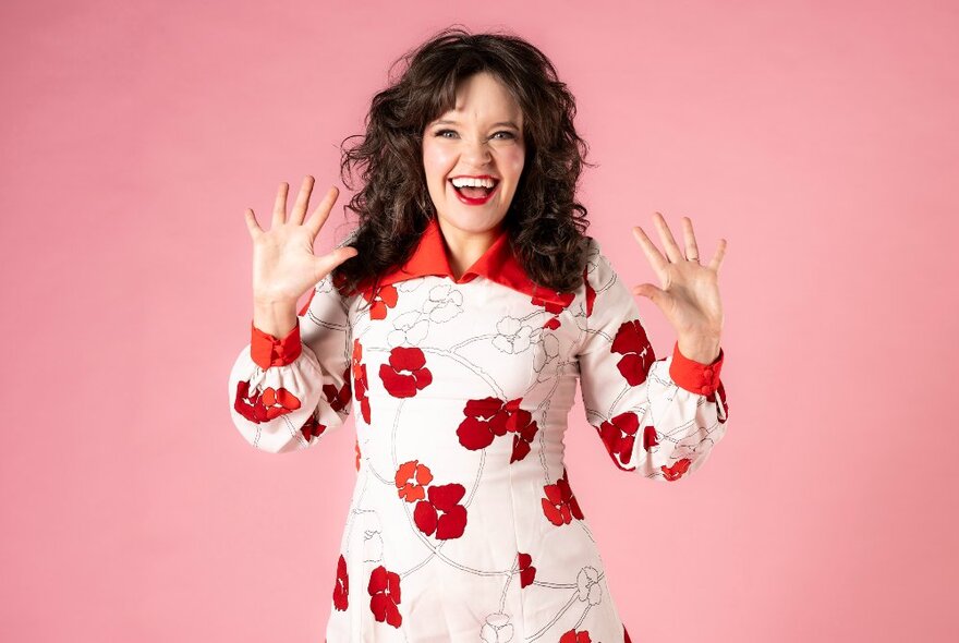 Singer and performer Alice Tovey in a white dress with a large red floral pattern on it, standing with their arms raised and palms facing outwards, in front of a pink backdrop.