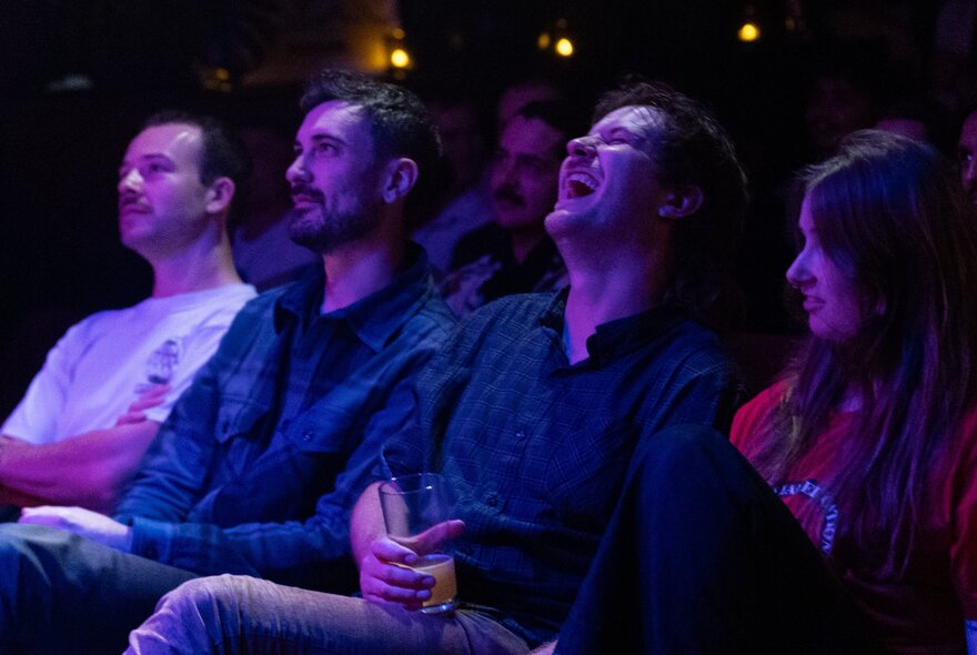 Four people seated in a row under dark purple lights, one person laughing.