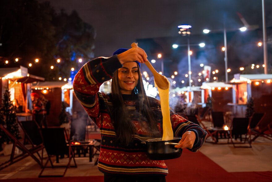 A person smiling and stretching a large amount of melted cheese from a fondue pot at an outdoor event at night.