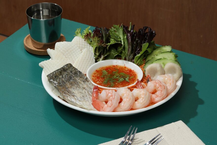 A dish of prawns and fish, ready to be cooked in a hot pot, with dipping sauce, and fresh salad greens accompanying, on a white plate on a table.