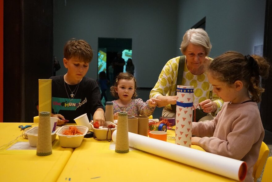 A group of three kids and an older woman at a yellow table all doing a craft activity with cardboard tubes. 