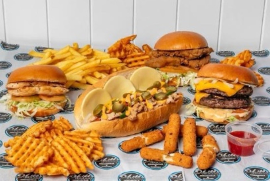 A table spread with Philly cheesesteak, three burgers, waffle fries, french fries, mozzerella sticks and dipping sauce. 