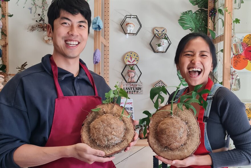 Two smiling class participants holding up their home-made kokedama balls of string topped with plants, in a workshop studio room with shelves draped with plants and crafts.