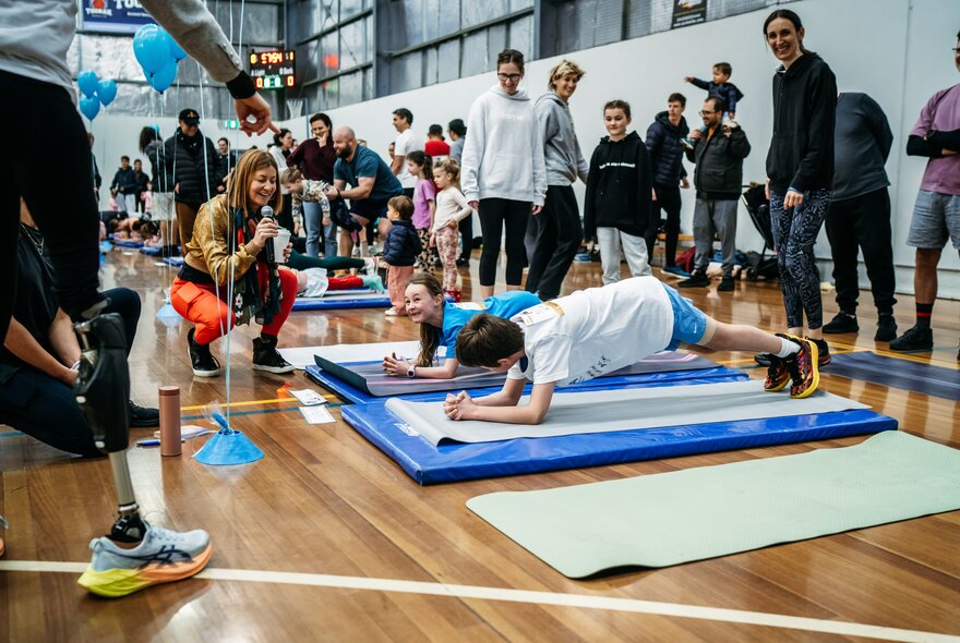 Two children doing planks on exercise mats in an indoor hall, surrounded by standing adults and spectators.