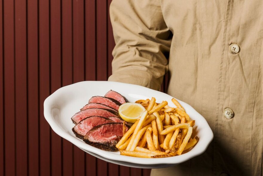 A waiter holding a white plate of steak frites, a classic French meal consisting of beefsteak paired with french fries.