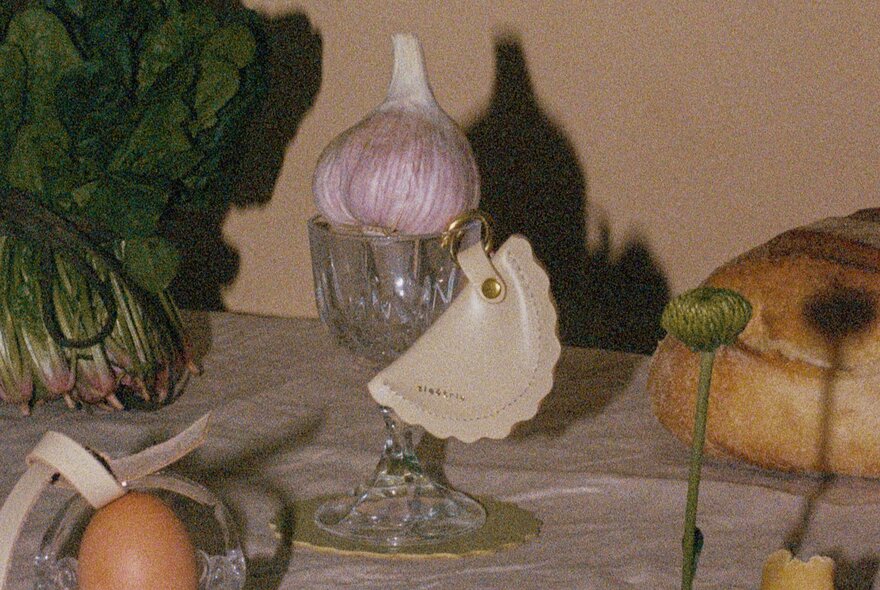 A still life arrangement of a bulb of garlic resting in a glass, a bunch of spinach, a loaf of bread and a small leather coin purse, arranged on a dining table.