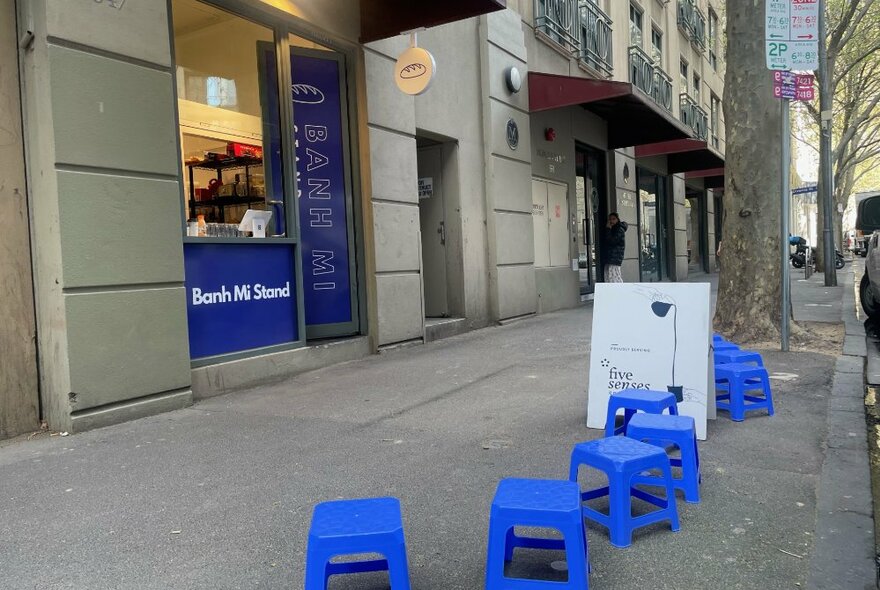 Small kiosk with a blue door, a window and blue stools out the front on the grey concrete path with a white, A-frame sign with incidental text behind.