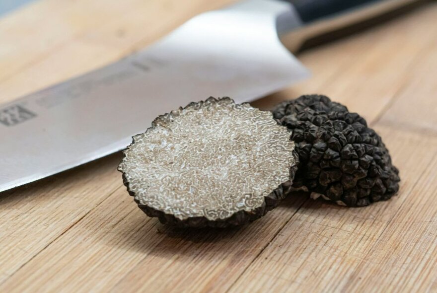 A black truffle, sliced in half with a sharp chef's knife behind it, both on a wooden chopping board.