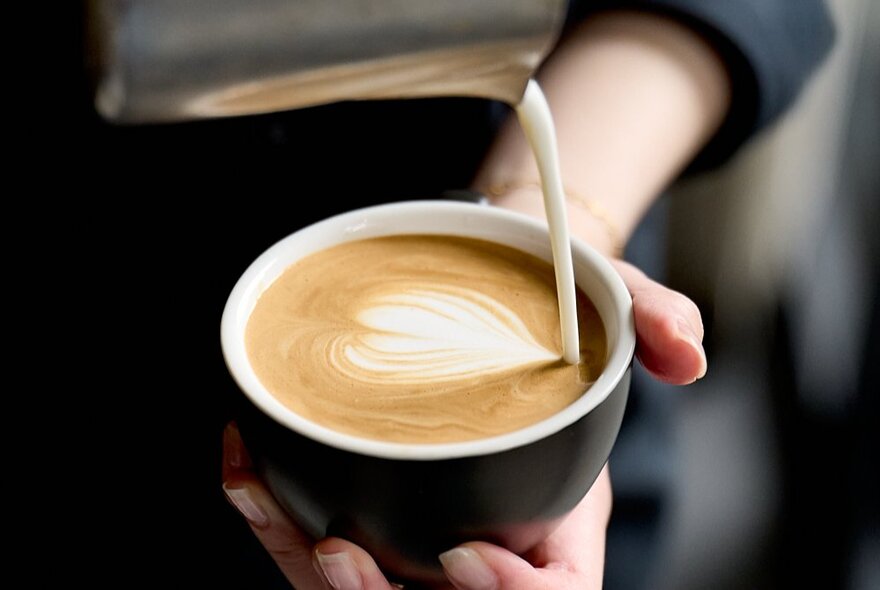 A barista pouring milk from a silver jug into a black ceramic cup of coffee.