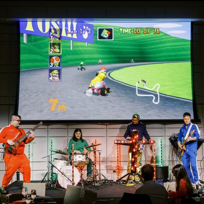 A live band performs on a stage in front of an audience, dressed in brightly coloured tracksuits while playing instruments. Behind them, a large projector screen displays a retro kart racing video game featuring a character driving on a track with green hills.