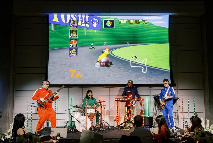 A live band performs on a stage in front of an audience, dressed in brightly coloured tracksuits while playing instruments. Behind them, a large projector screen displays a retro kart racing video game featuring a character driving on a track with green hills.