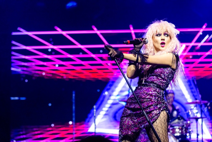 A female singer wearing a purple sequinned dress, singing on stage with neon pink stage lighting behind.