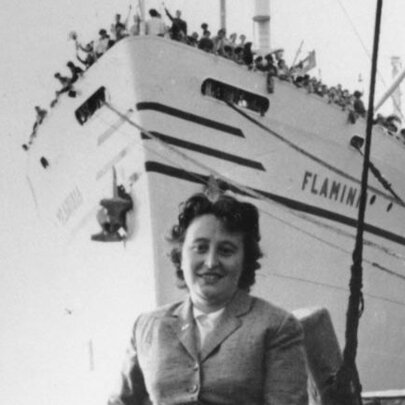 Italian woman standing in front of a ship with passengers crowding the prow deck.